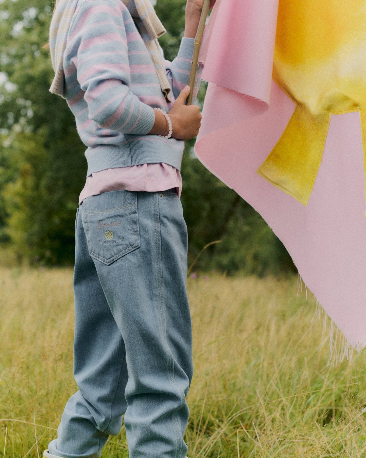 Child wearing light blue denim jeans with relaxed fit and pastel striped sweater holding a pink and yellow flag outdoors