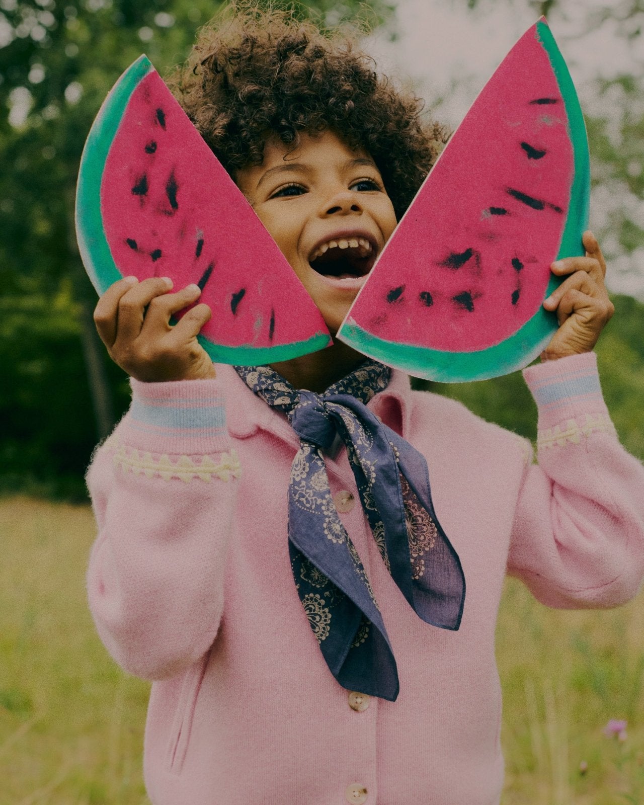 Child wearing a pink cardigan and blue patterned scarf holding two large watermelon slice props outdoors