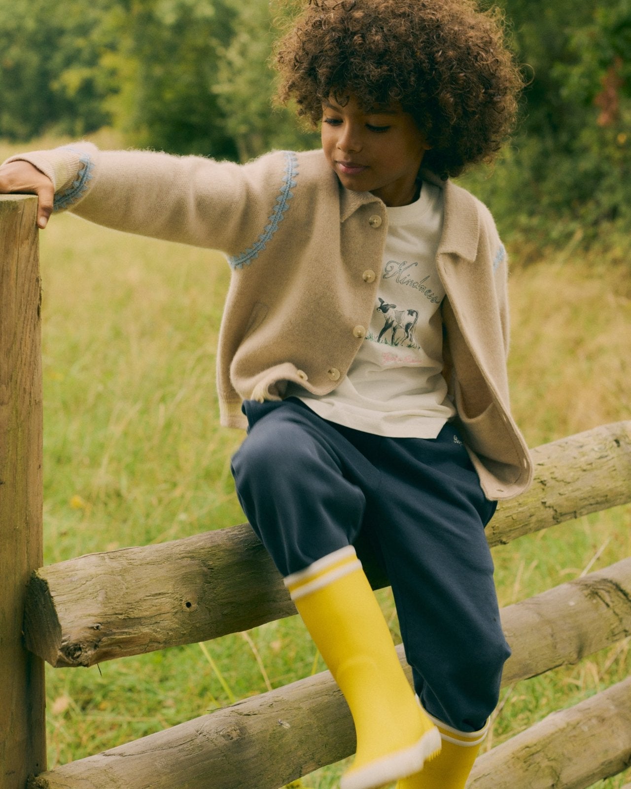Children's beige cardigan with blue embroidery, paired with navy pants and yellow rain boots, outdoor countryside setting