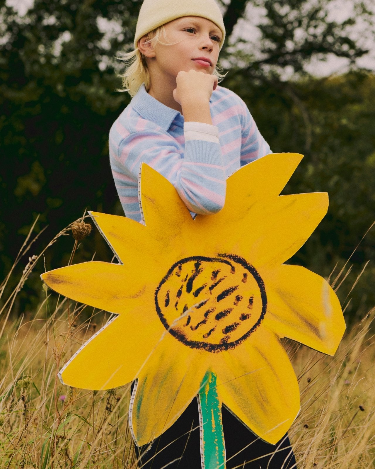 Child in pastel striped sweater and cream beanie holding large yellow sunflower prop outdoors in nature setting