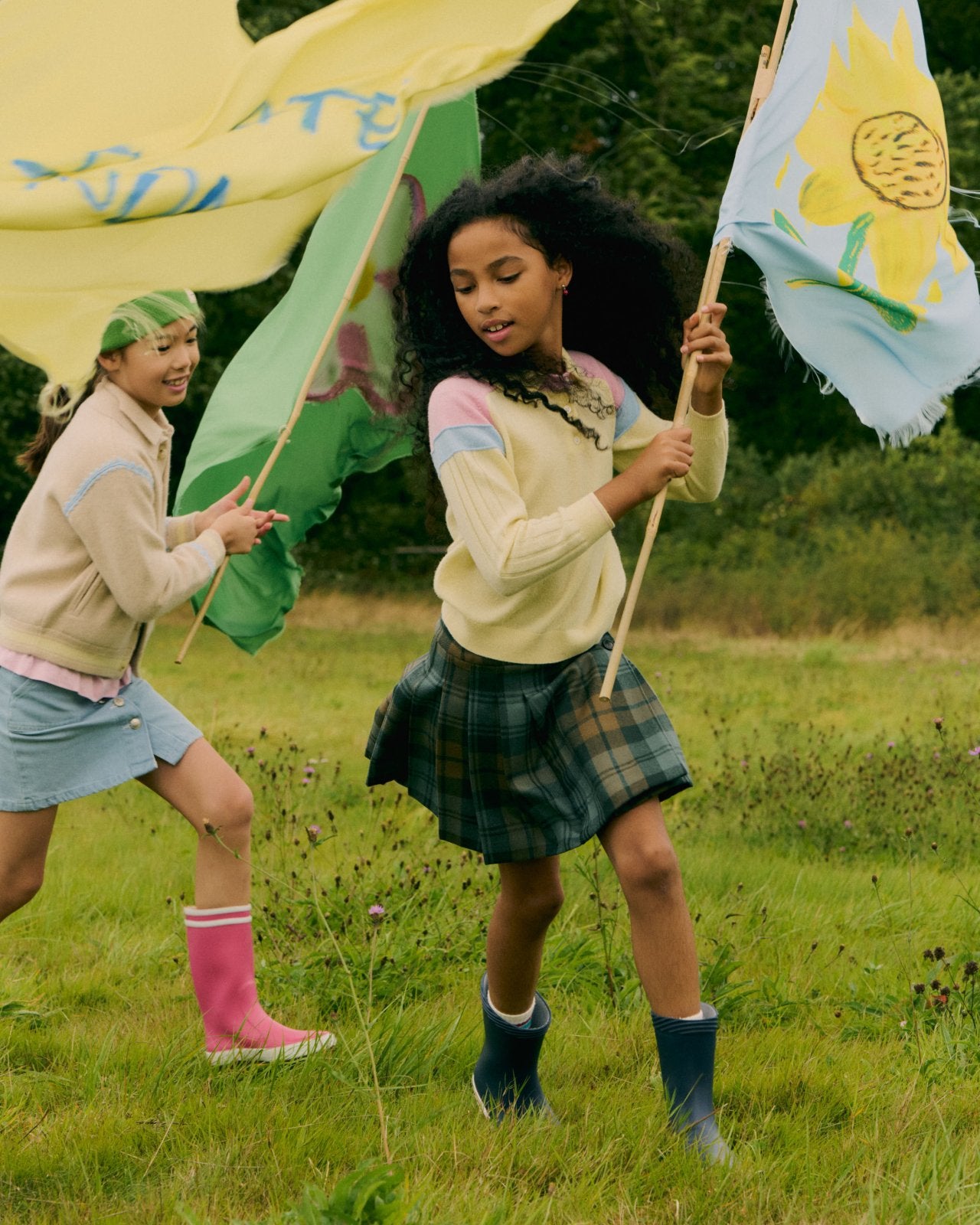 Two girls playing outdoors holding colorful flags, one wearing a pastel sweater with plaid skirt and rain boots