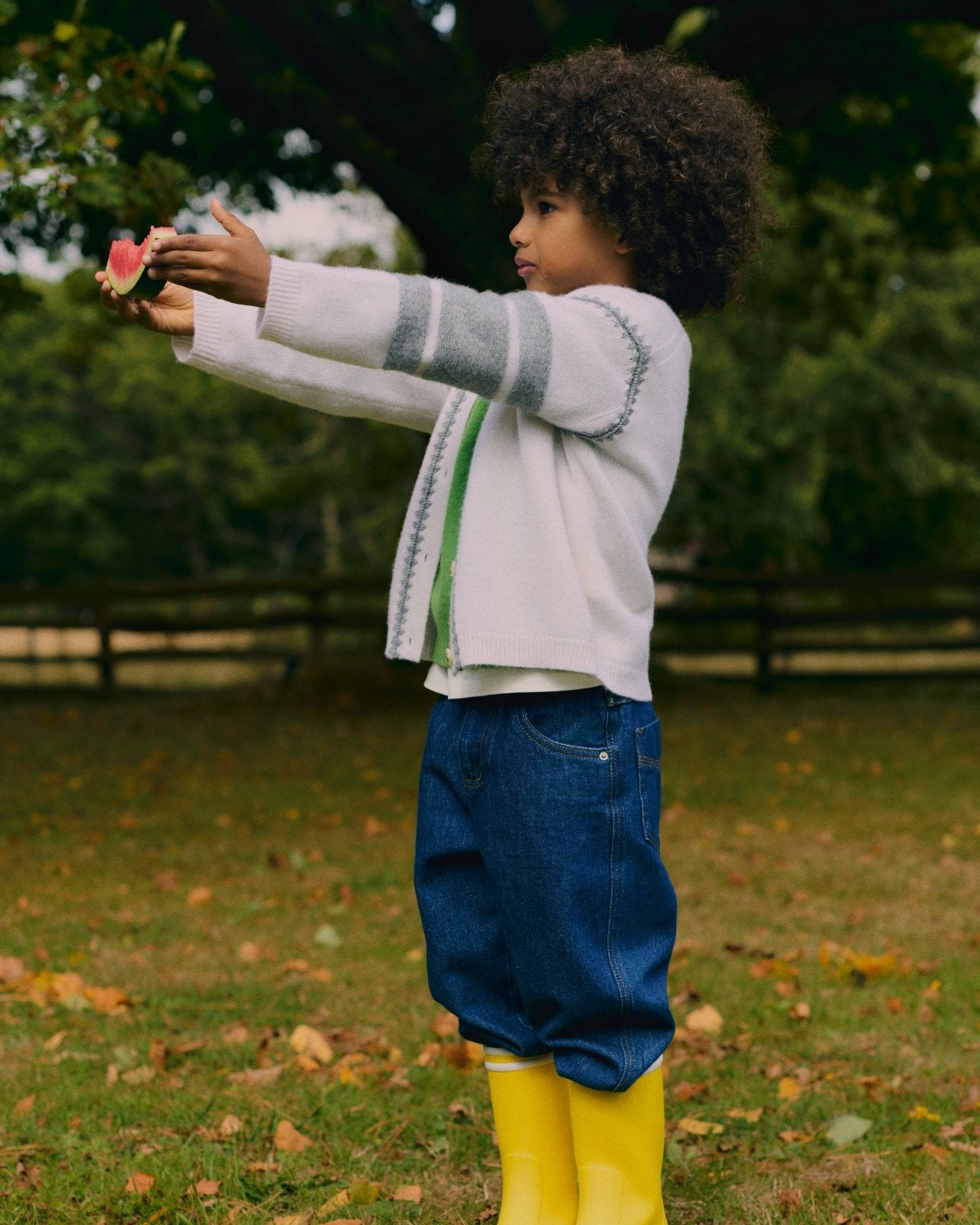 Child wearing white cardigan with gray stripes, blue jeans, and yellow rain boots, holding a slice of watermelon outdoors.