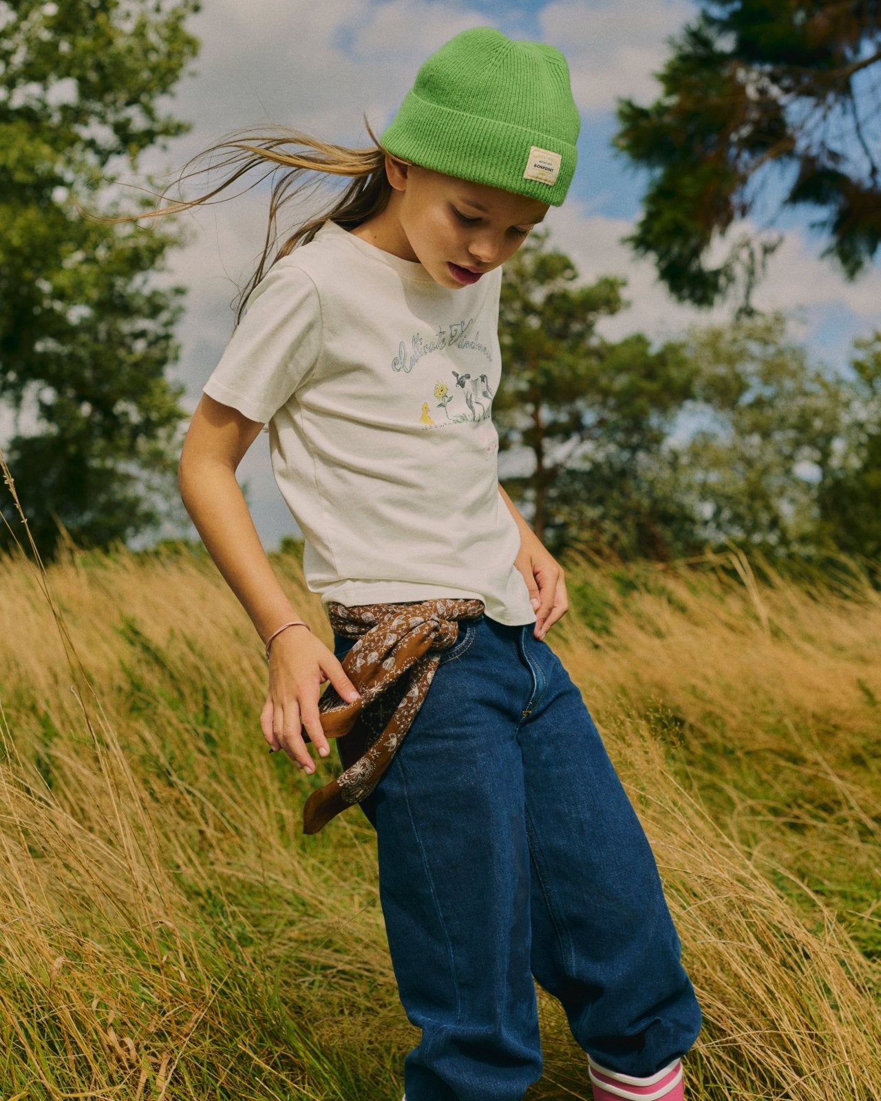Child wearing white graphic tee and blue jeans with brown patterned scarf belt and green knit beanie outdoors in field