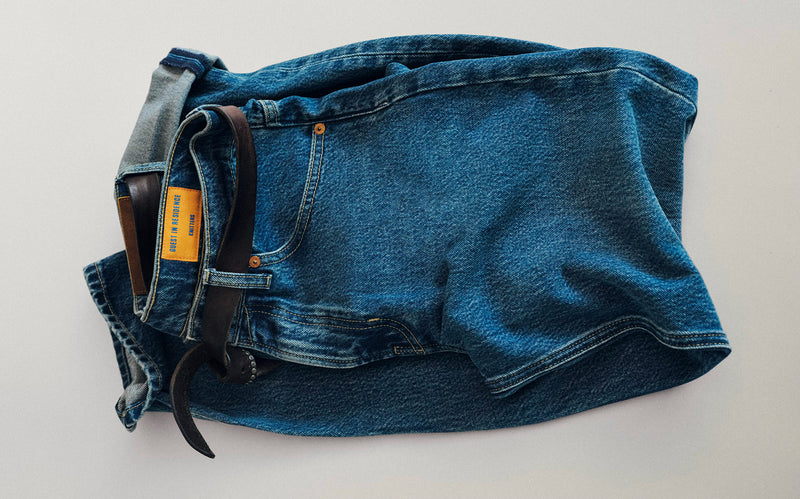Folded blue denim jeans with visible front pockets, belt loops, stitching, waistband, and a small tan label against a light background