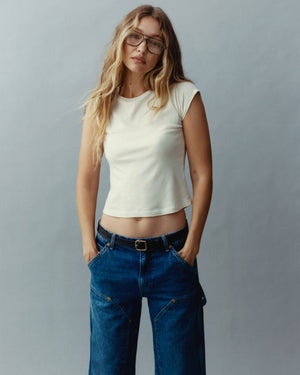 Woman wearing glasses and a white sleeveless top with loose blue jeans and a belt standing against a gray studio background