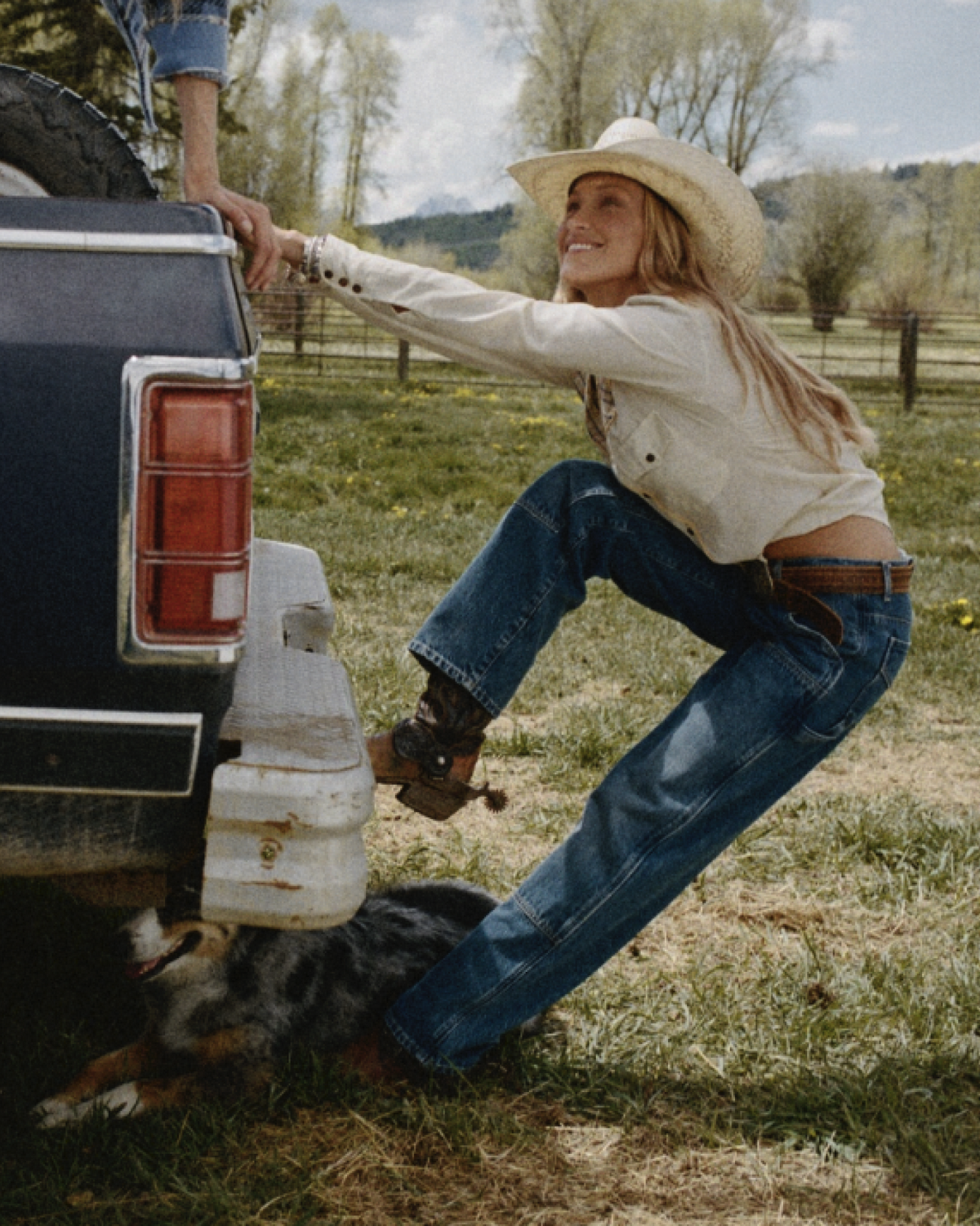 Woman in a cowboy hat and cream sweater pulls on the tailgate of a vintage truck while a dog lies beneath it in a grassy field