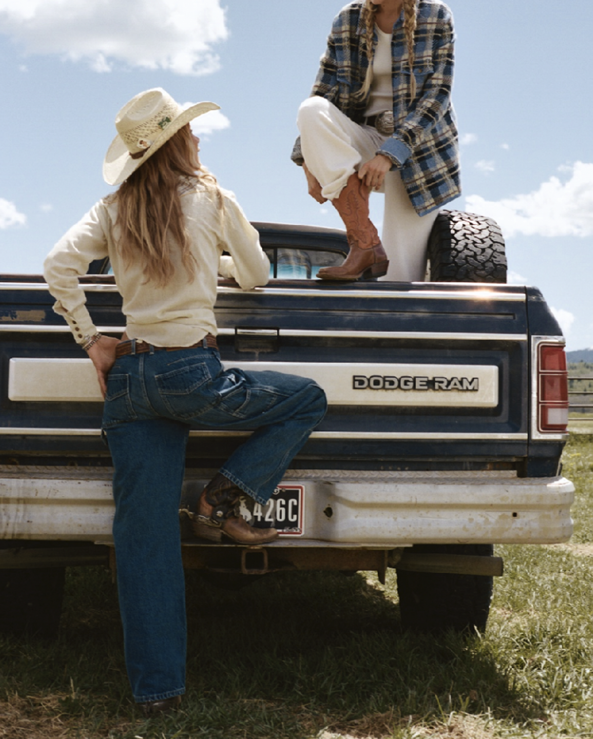 Two people pose beside a vintage Dodge Ram pickup truck in a grassy field under a blue sky one wearing a cowboy hat and the other wearing a plaid jacket