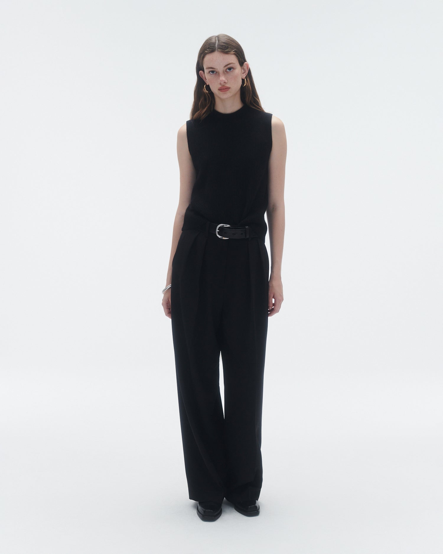 Full body of female model wearing a black cashmere vest tucked into black trousers with a belt