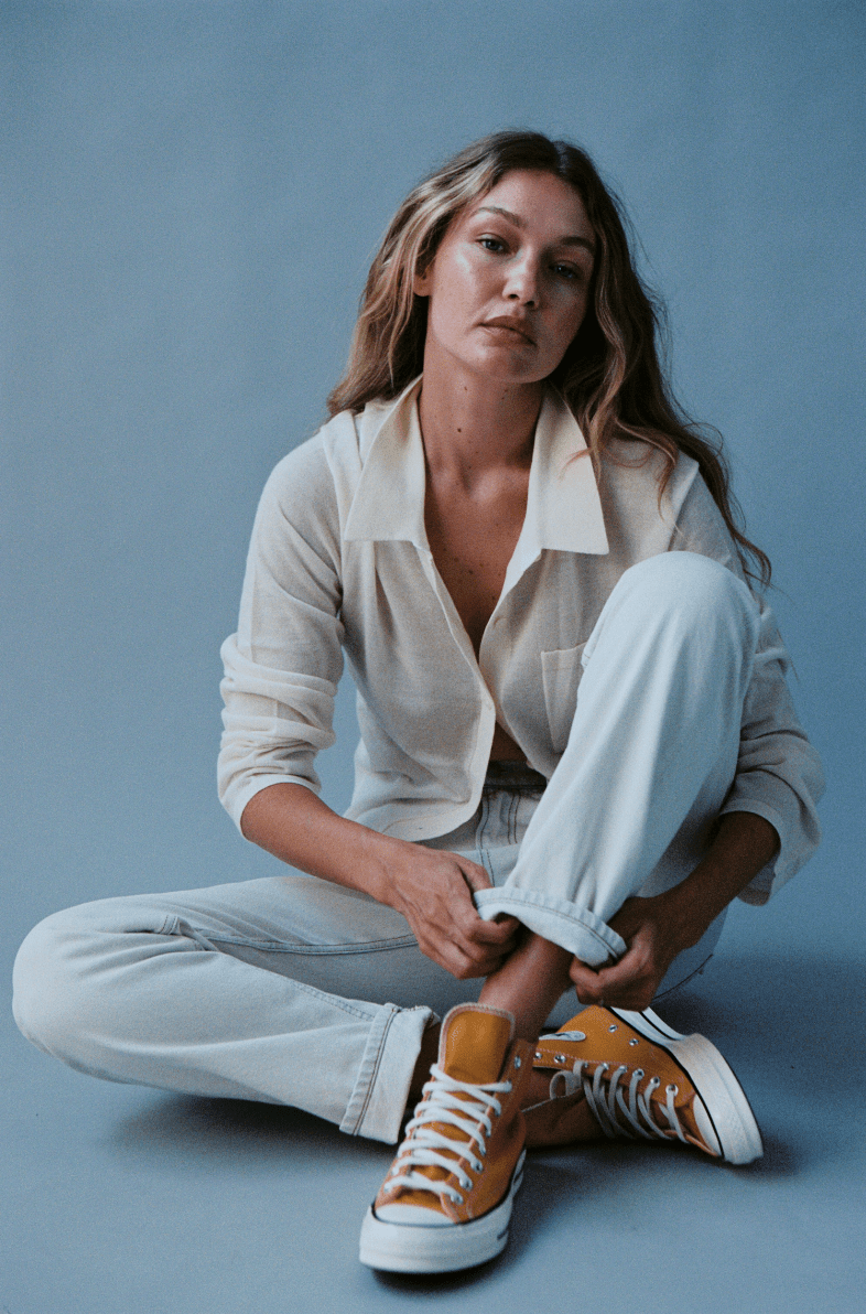 EVERYDAY model seated in a light button up shirt and relaxed trousers with rolled cuffs and contrasting high top sneakers against a blue studio backdrop