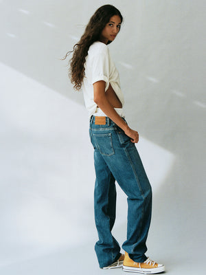 Woman with long curly hair wearing a white T shirt, relaxed blue jeans, and white sneakers while standing in profile against a softly lit studio wall