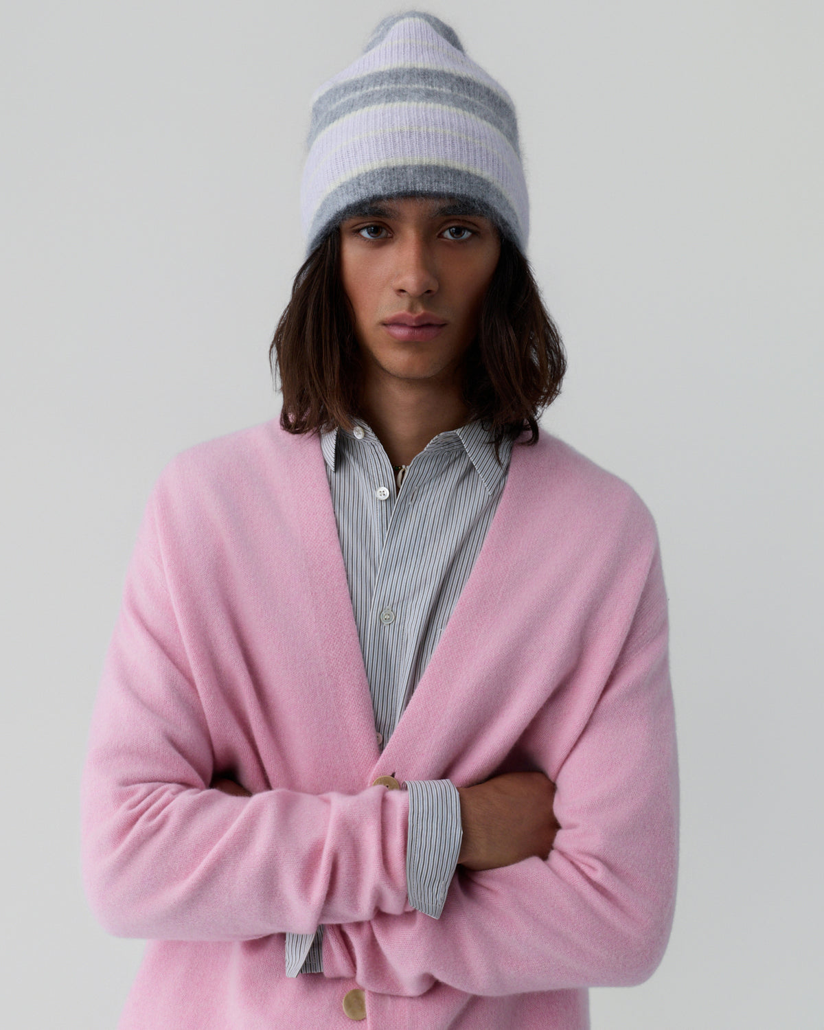 Person with shoulder-length dark hair wearing a soft pink cardigan over a gray and white striped button-up shirt and a knitted beanie with pale purple and gray stripes against a neutral background.