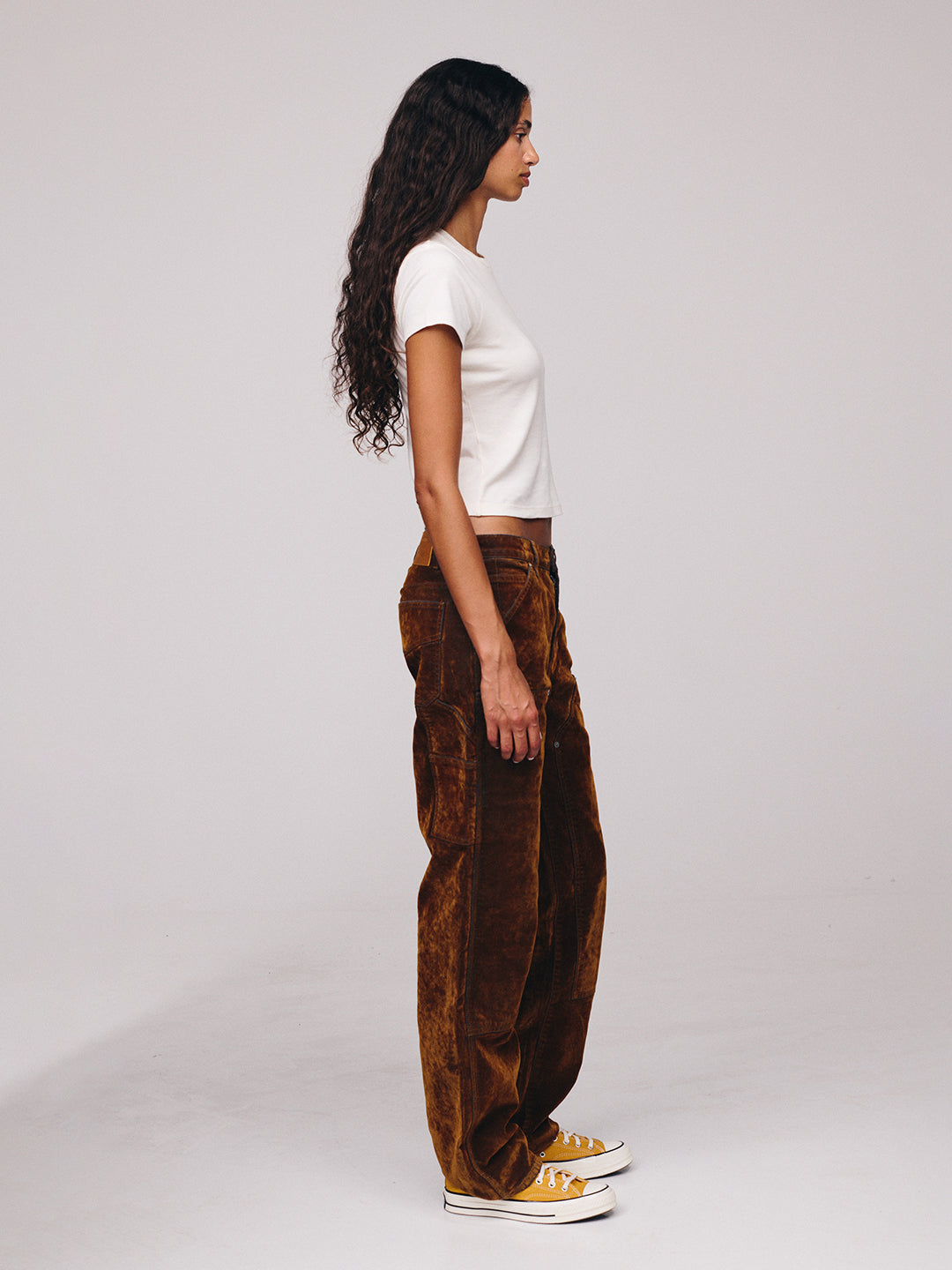Side profile of a woman with long curly hair wearing a white T shirt, brown patterned loose fit trousers, and white sneakers in a studio setting