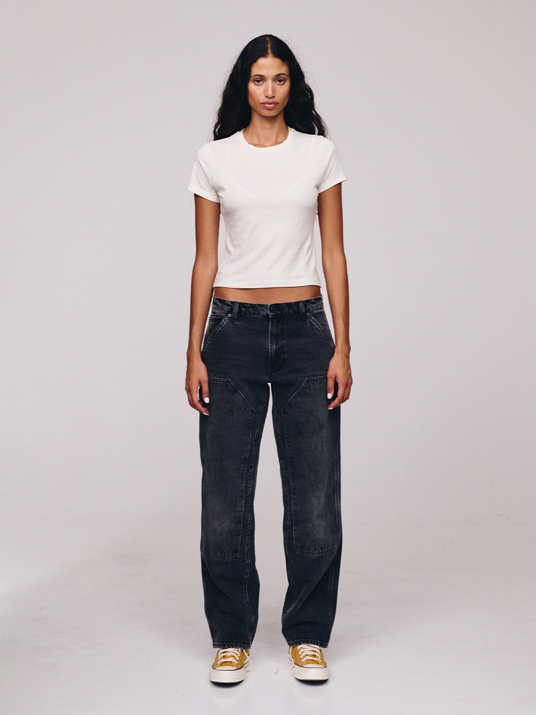 Full length portrait of a woman with long dark hair wearing a white fitted T shirt dark wide leg jeans and orange and white sneakers against a plain studio backdrop