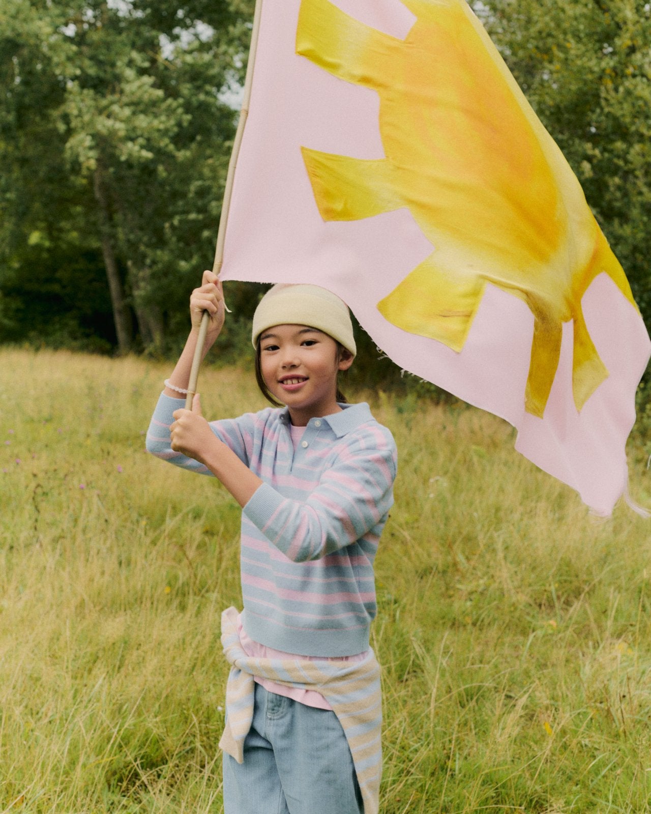 Child wearing a light blue and pink striped long-sleeve polo with a yellow beanie, holding a pink flag with a yellow sun design outdoors