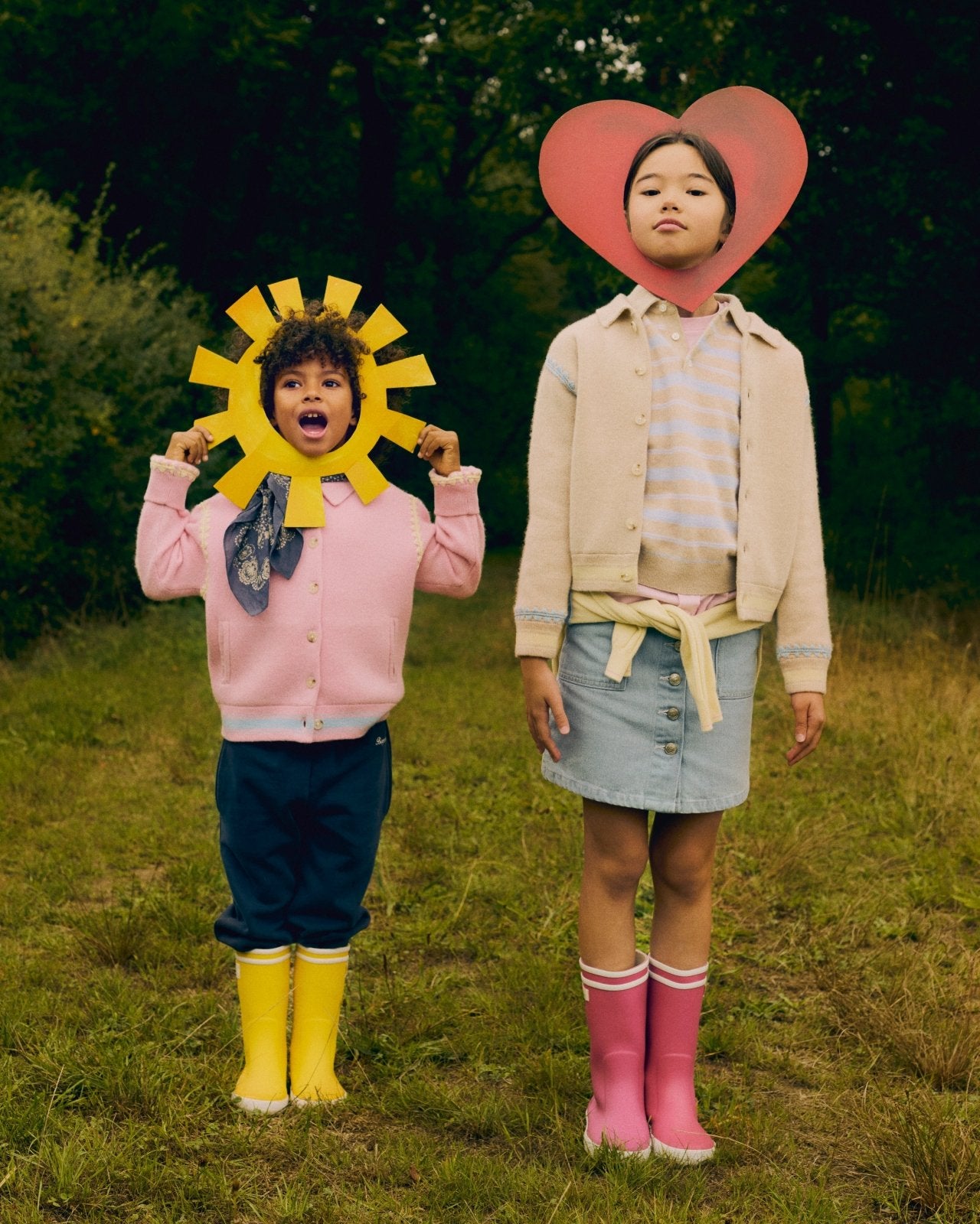 Two children outdoors wearing pastel sweaters and colorful rain boots with playful sun and heart headpieces