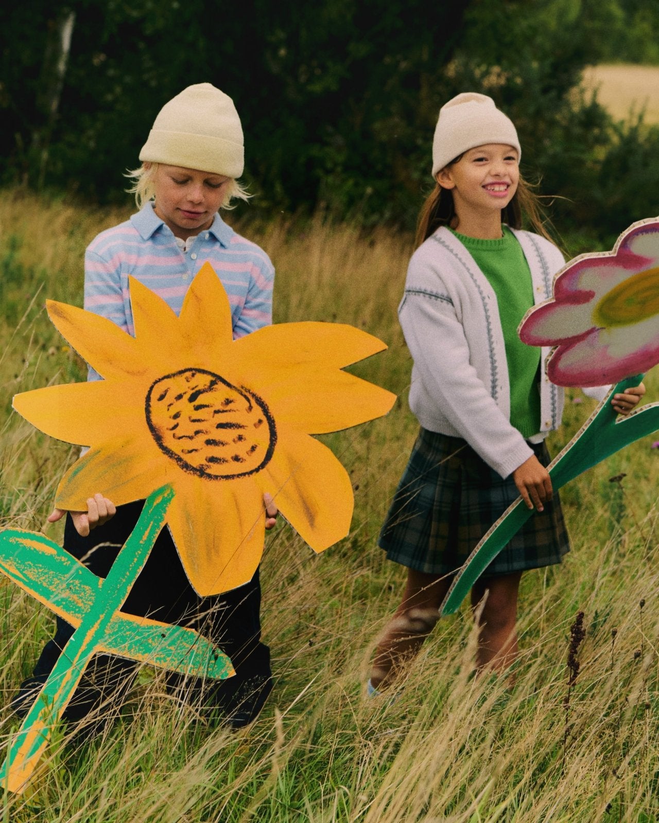 Two children wearing knit hats and layered outfits holding large colorful flower cutouts outdoors in a grassy field