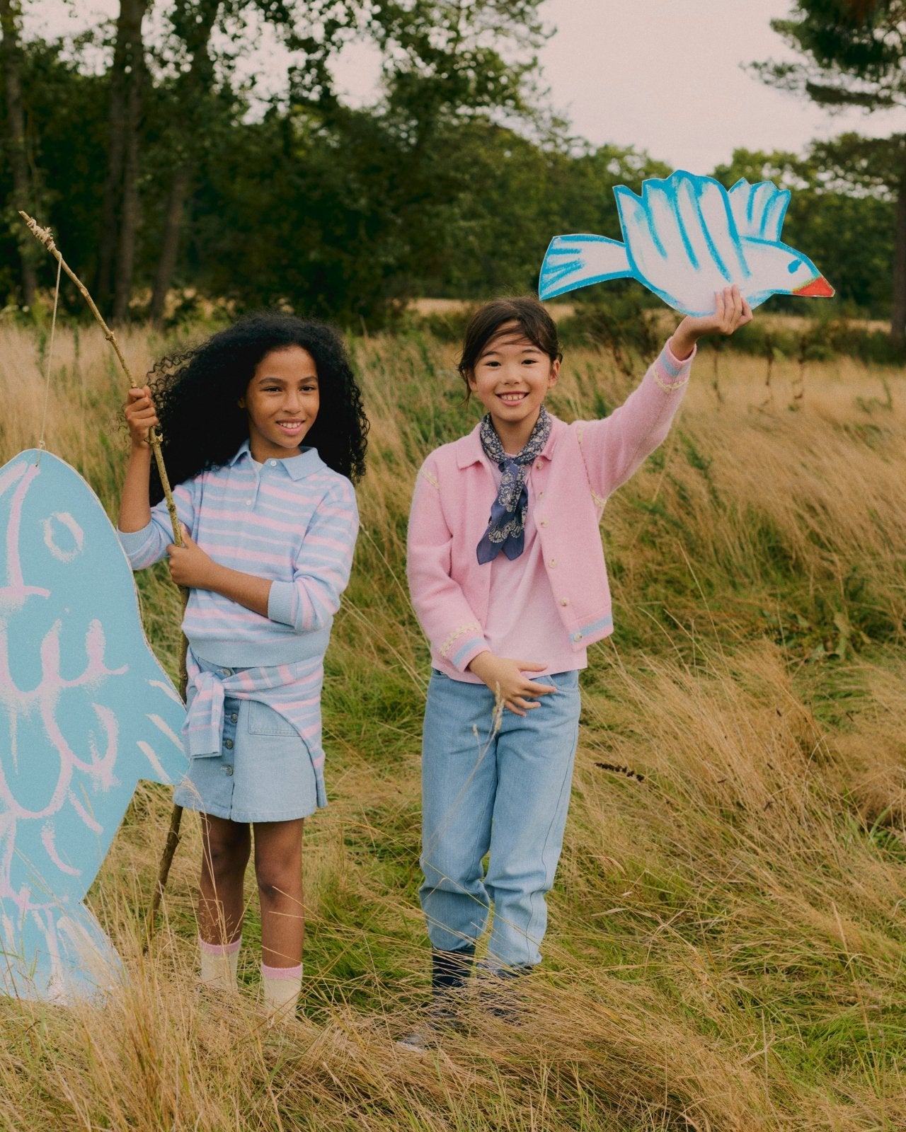 Two children outdoors holding large blue bird cutouts, wearing casual pastel sweaters and denim in grassy field setting