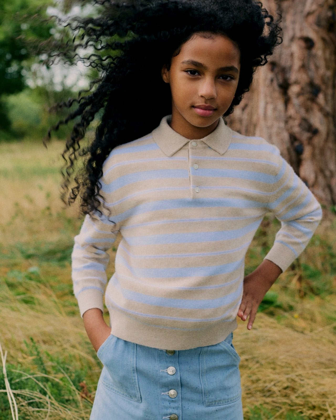 Children's lightweight beige and white striped polo sweater with ribbed cuffs and collar, paired with button-front denim skirt outdoors.