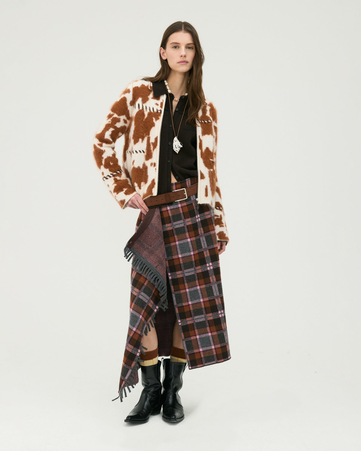 Woman wearing a brown and cream patterned cardigan, black top, plaid wrap skirt with fringe, layered scarf, belt, and black boots.