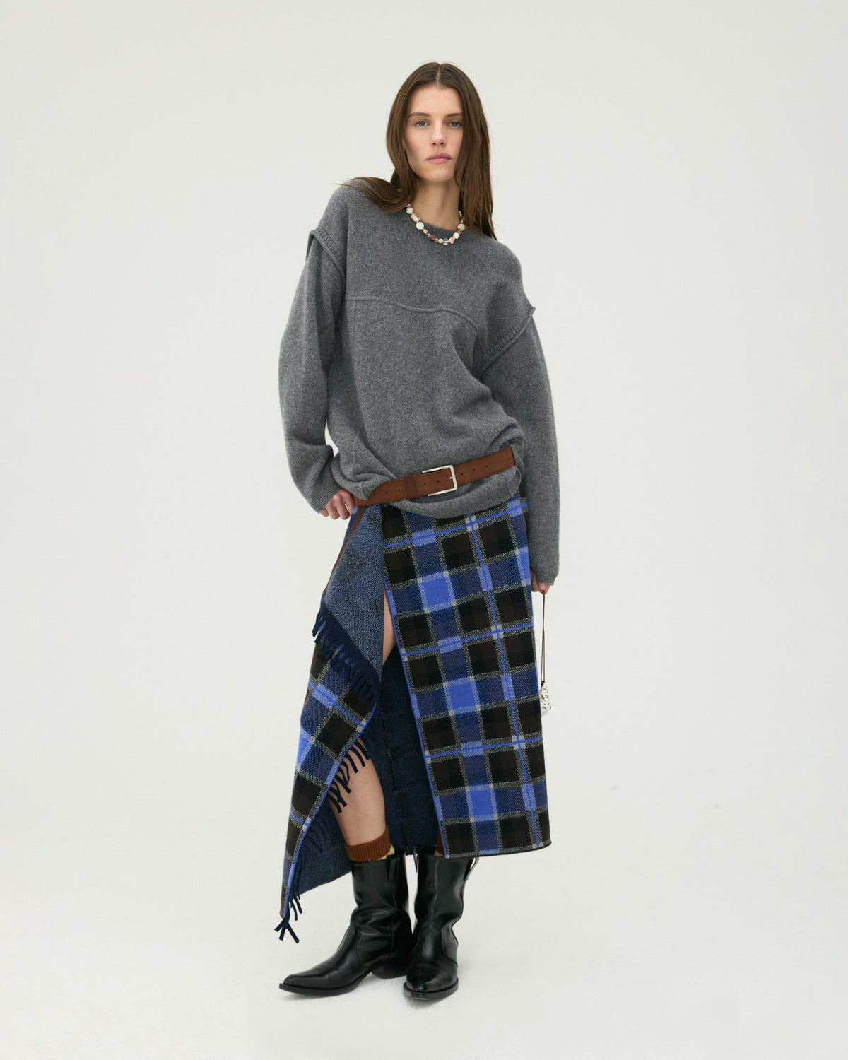 Woman wearing a charcoal gray sweater with a blue plaid wrap skirt and black knee high boots against a light neutral studio background