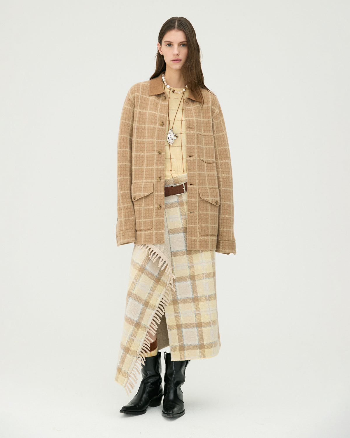 Model wearing a tan plaid cardigan with a long fringed scarf, cream top, coordinated plaid skirt, and black knee high boots against a light background