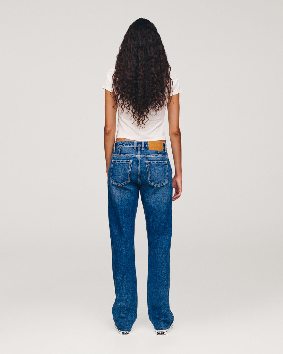 Rear view of a woman with long curly hair wearing a white T shirt and blue wide leg jeans against a plain light background