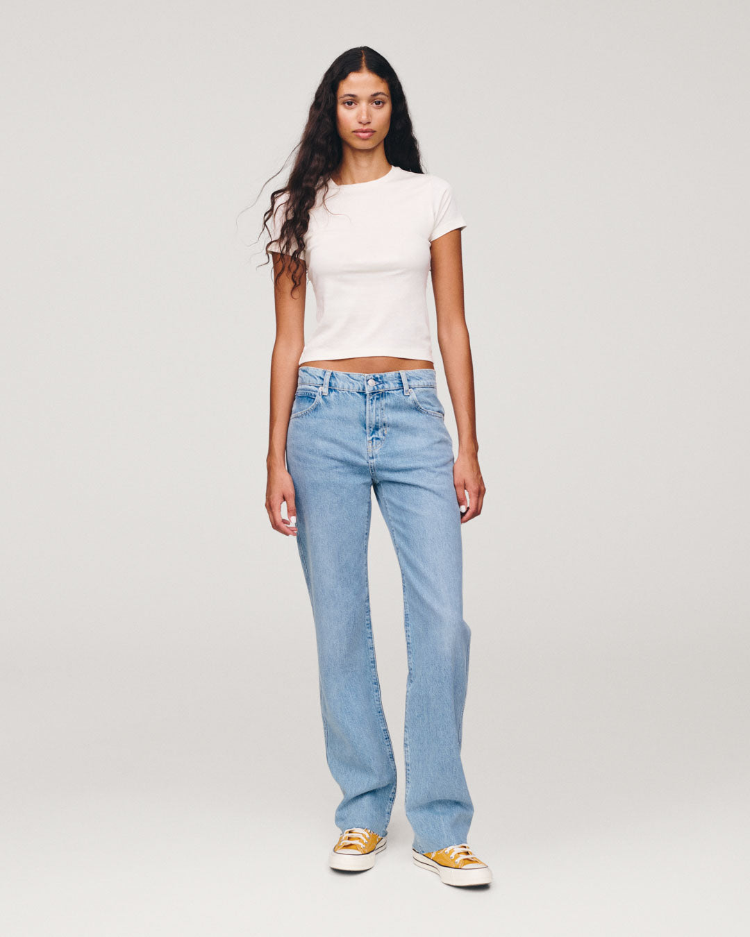 Full length view of a woman wearing a white cropped T shirt light blue wide leg jeans and orange and white sneakers against a plain light gray background