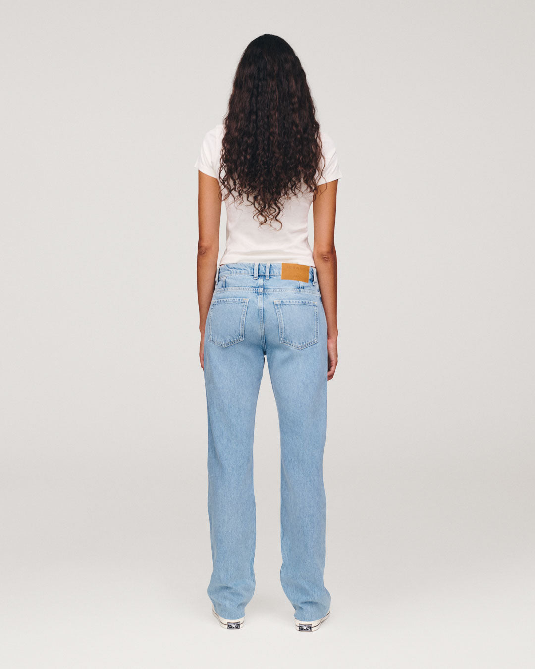 Back view of a woman with long curly hair wearing a white T shirt and light blue high waisted jeans against a plain light background