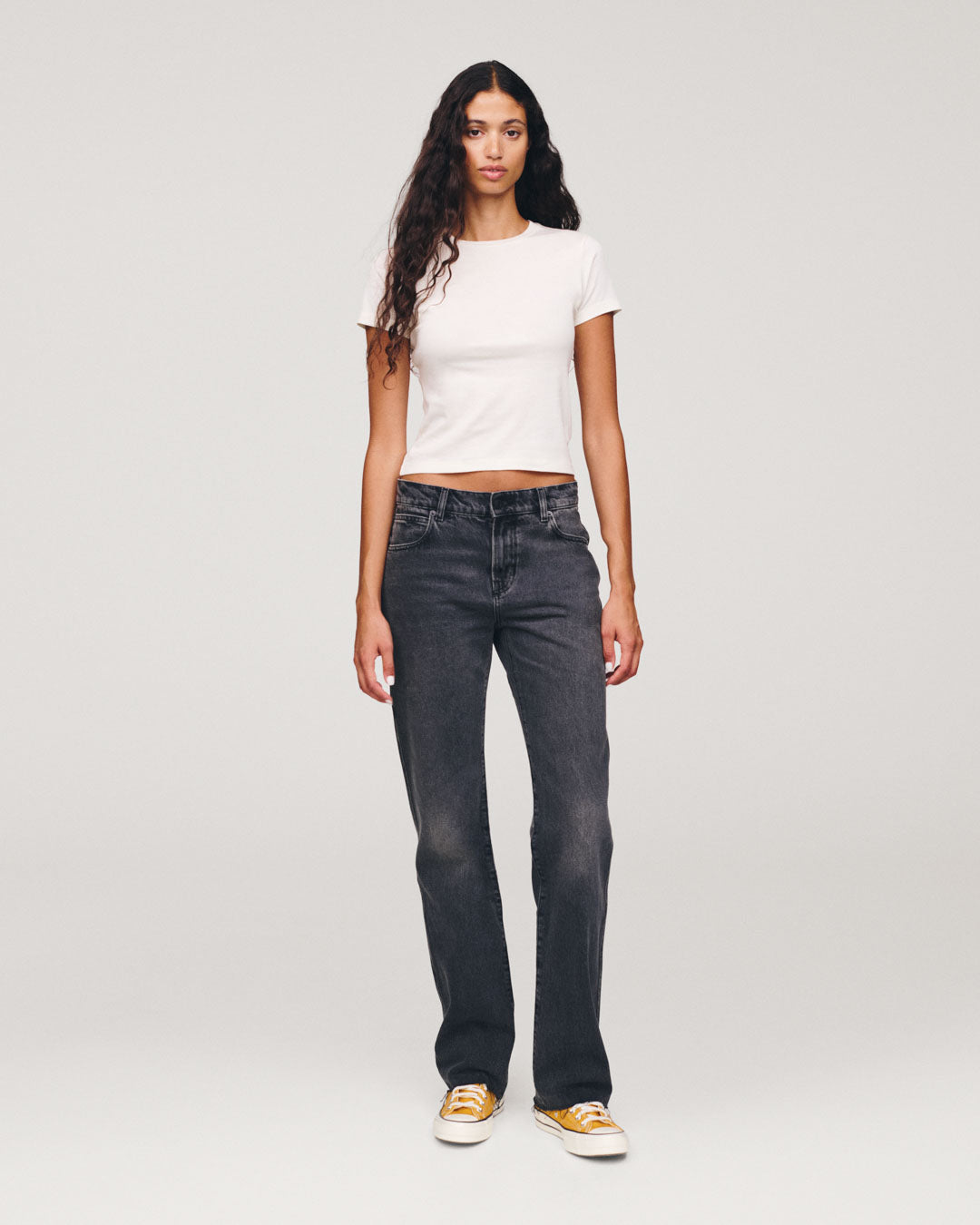 Woman with long dark hair wearing a white short sleeve top dark gray jeans and yellow and white sneakers against a plain light background