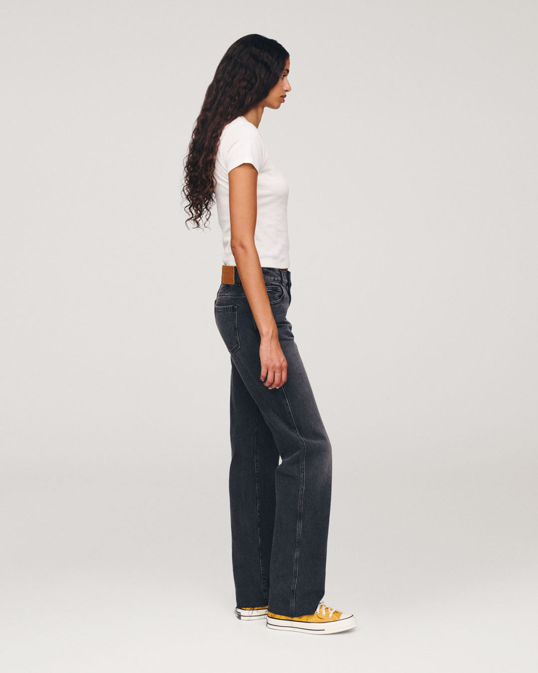 Side profile of a woman with long dark hair wearing a white T shirt dark wide leg jeans and yellow sneakers against a light gray background