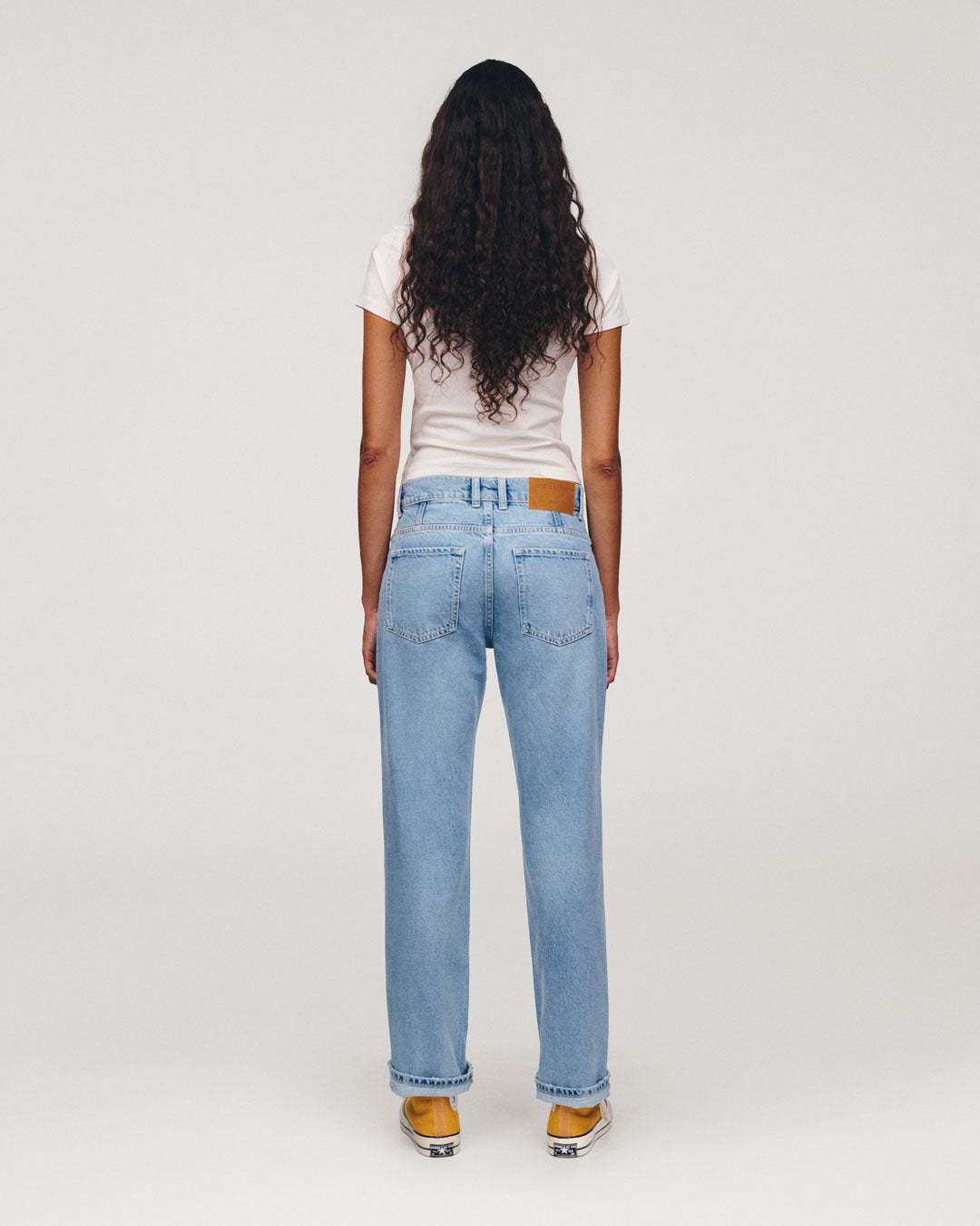 Rear view of a person with long curly hair wearing a white T shirt, light blue straight leg jeans, and yellow sneakers against a plain light background