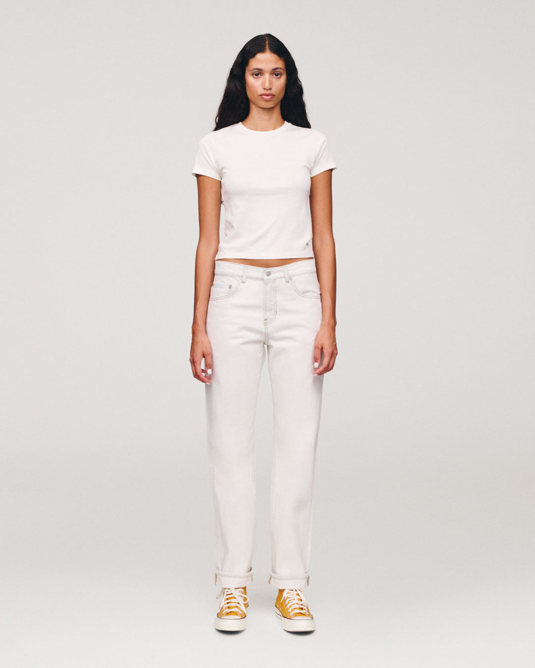 Full-length portrait of a woman wearing a white fitted T-shirt and white jeans with yellow and white sneakers against a plain light gray background