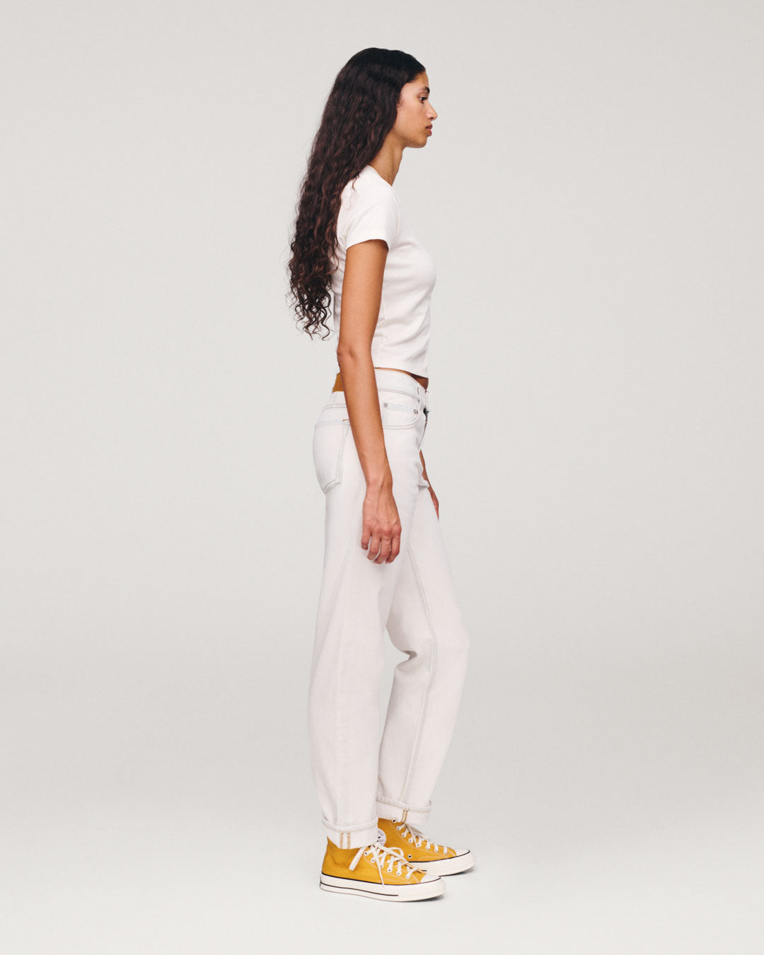 Side profile of a woman wearing a white T shirt and white jeans with bright yellow high top sneakers against a plain light gray studio background