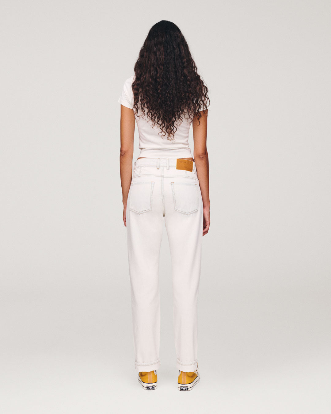 Back view of a woman with long curly hair wearing a white T shirt white jeans and yellow shoes against a light gray studio background