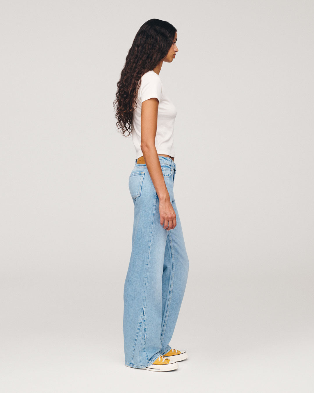 Side view of a woman with long curly hair wearing a white T shirt, wide leg light blue jeans, and yellow sneakers against a plain light gray background