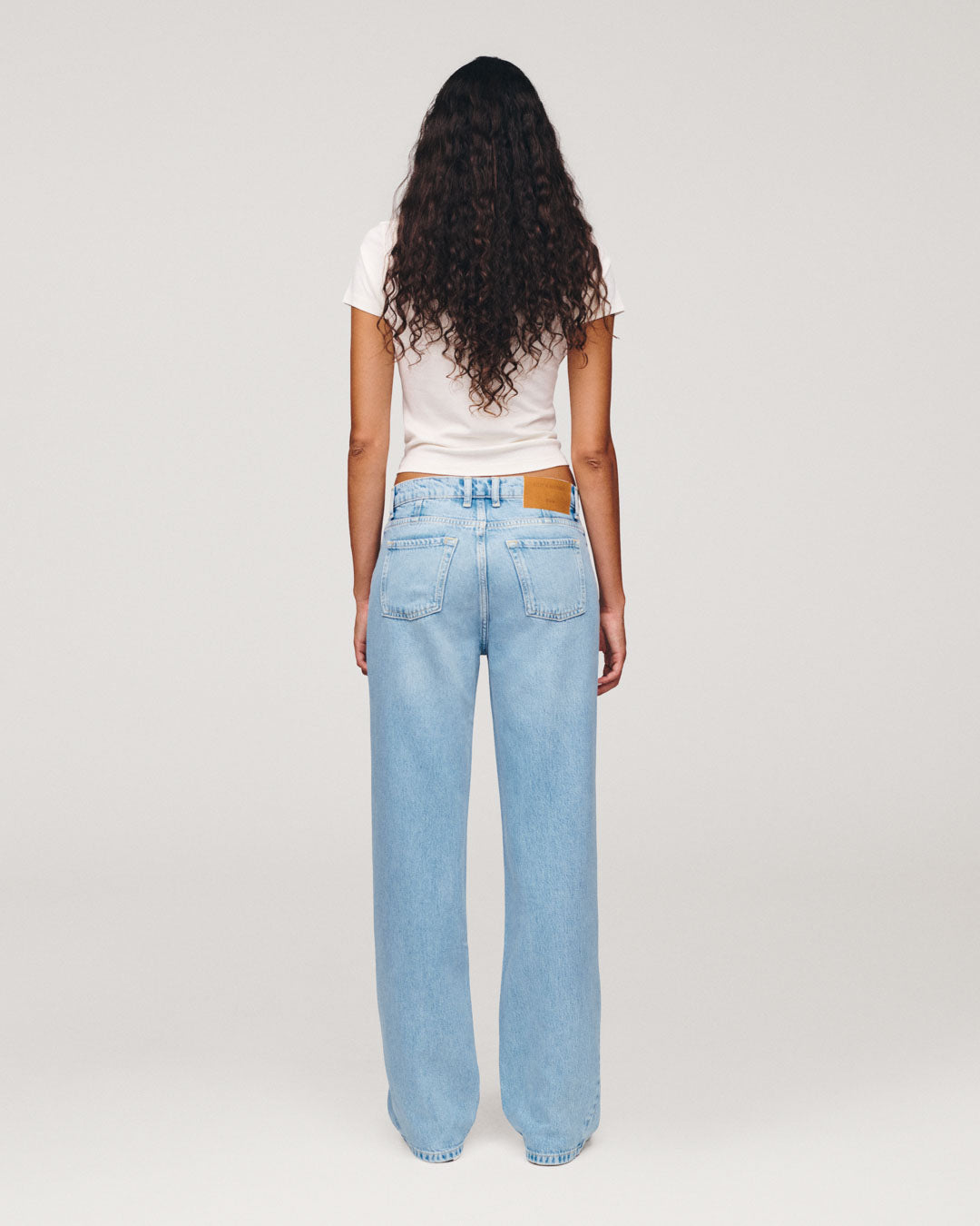 Back view of a model with long curly hair wearing a white T-shirt and loose light blue jeans against a plain gray studio background.