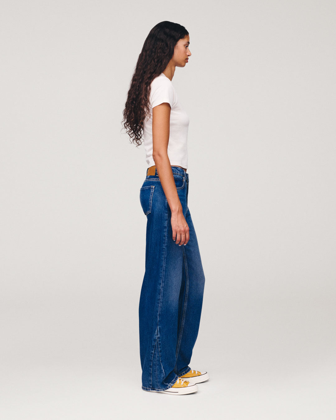 Side profile of a woman wearing a white T shirt with wide leg blue jeans and white sneakers against a light gray studio background