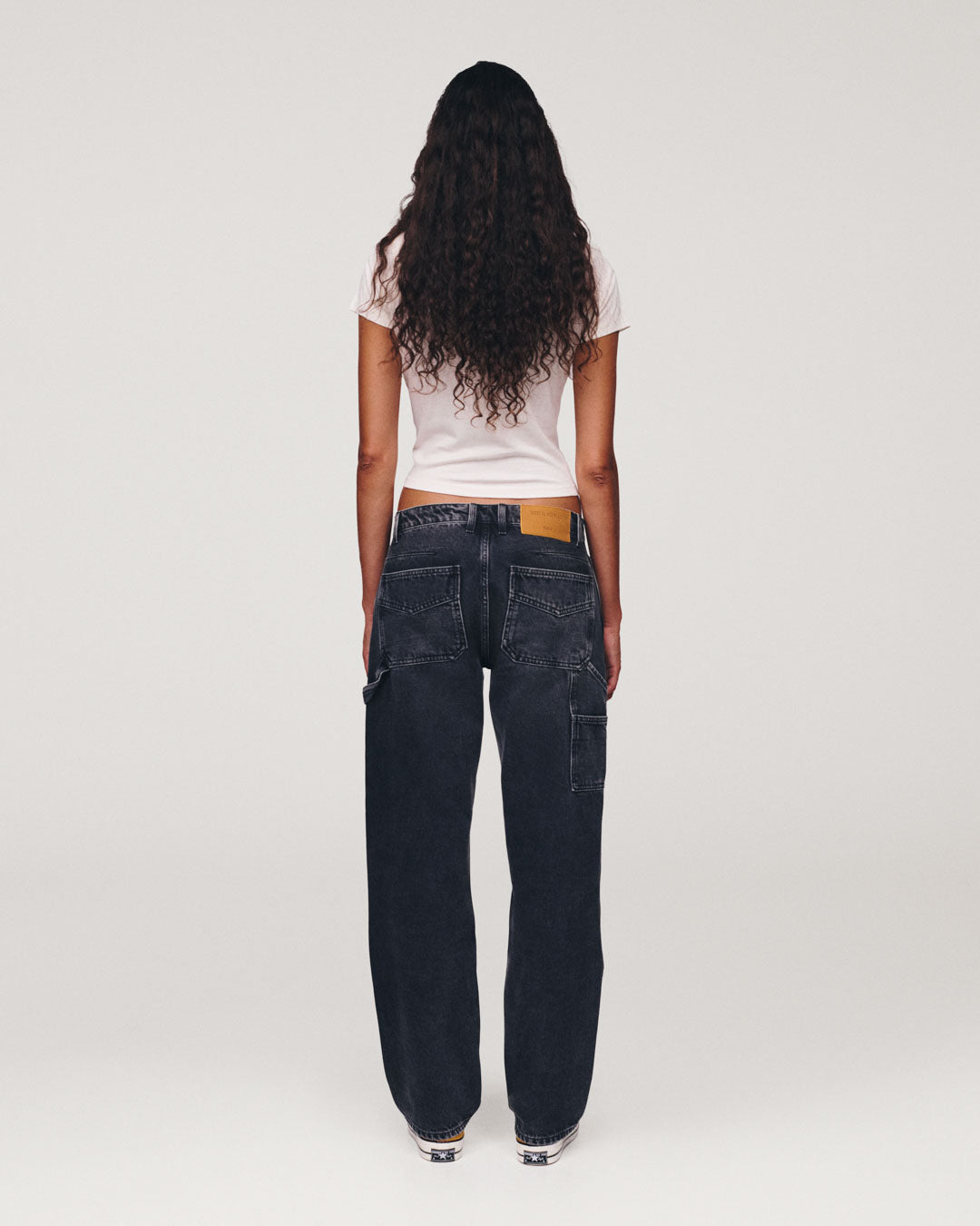 Woman standing with her back to the camera wearing a white T shirt and dark wide leg jeans against a plain light background
