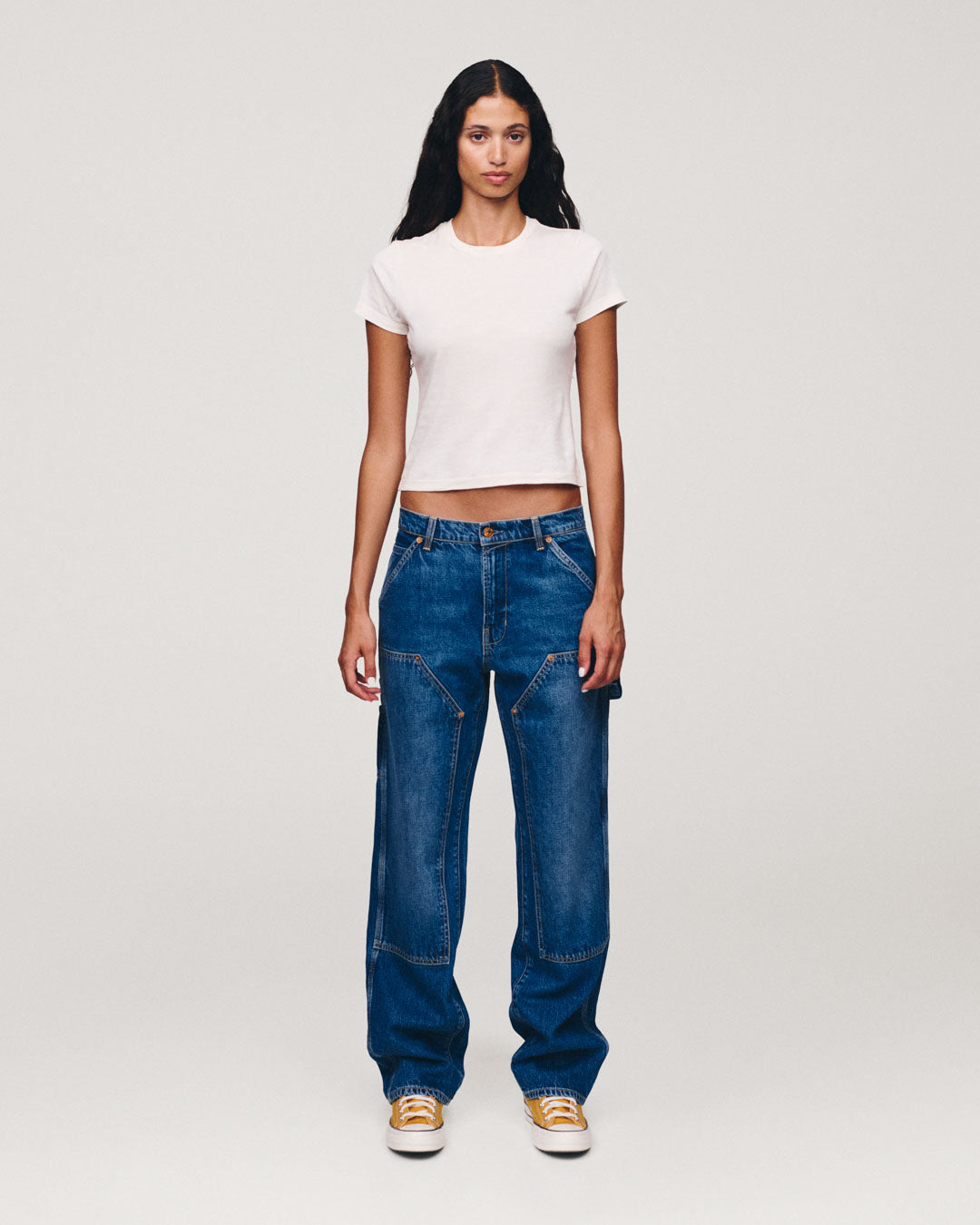 Full-length portrait of a woman wearing a white fitted T-shirt, relaxed blue jeans, and white sneakers with yellow accents against a light gray background