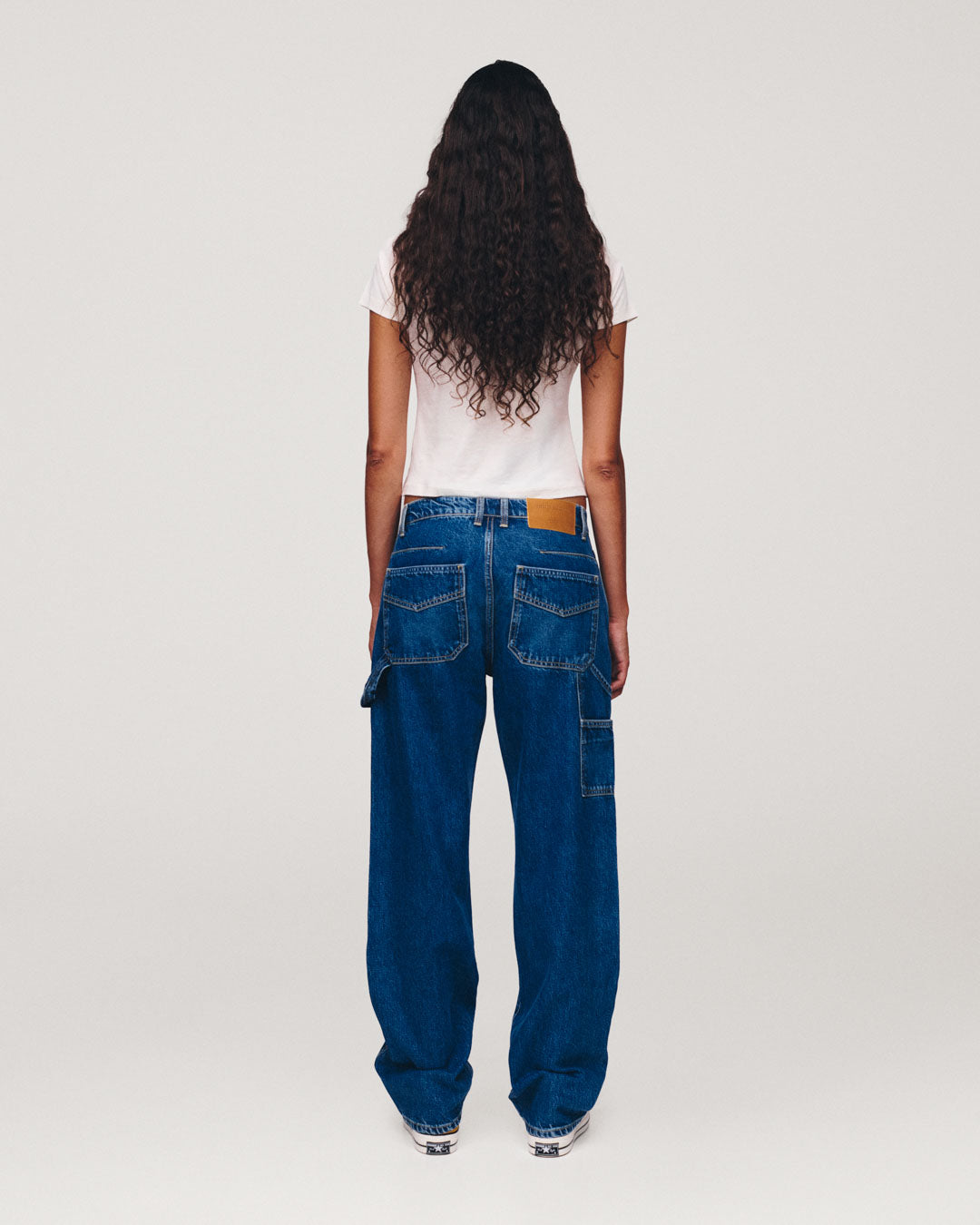 Back view of a person with long curly hair wearing a white T shirt and loose blue cargo jeans with multiple pockets against a plain light background