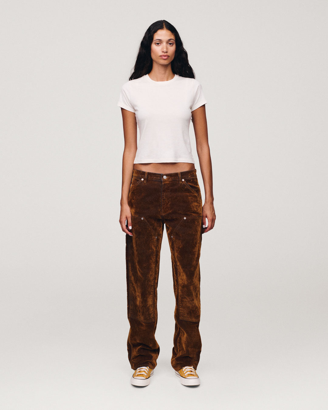 Woman standing in a studio wearing a fitted white short sleeve shirt brown textured trousers and white sneakers with orange accents