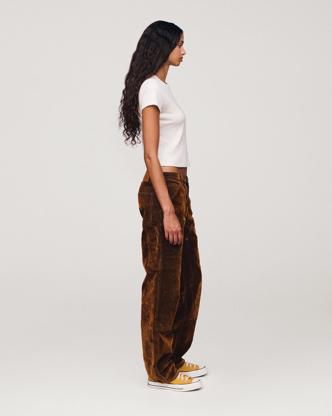 Side profile of a woman with long curly hair wearing a fitted white T shirt brown pants and white sneakers against a plain light gray background
