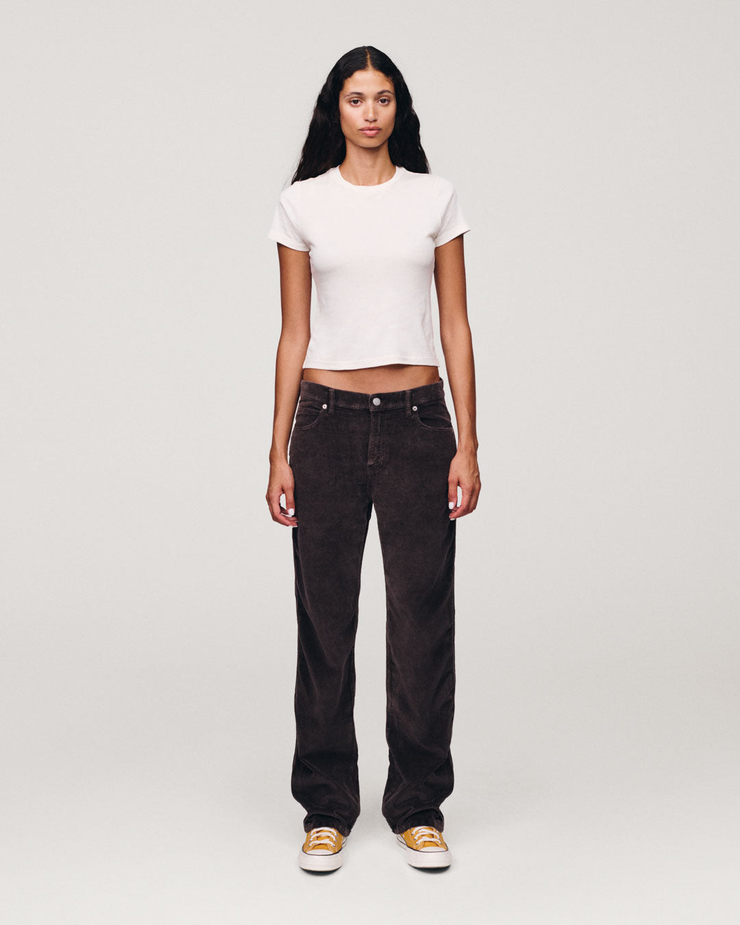 Model standing in a studio wearing a fitted white short sleeve top with dark brown jeans and yellow and white sneakers against a light gray background