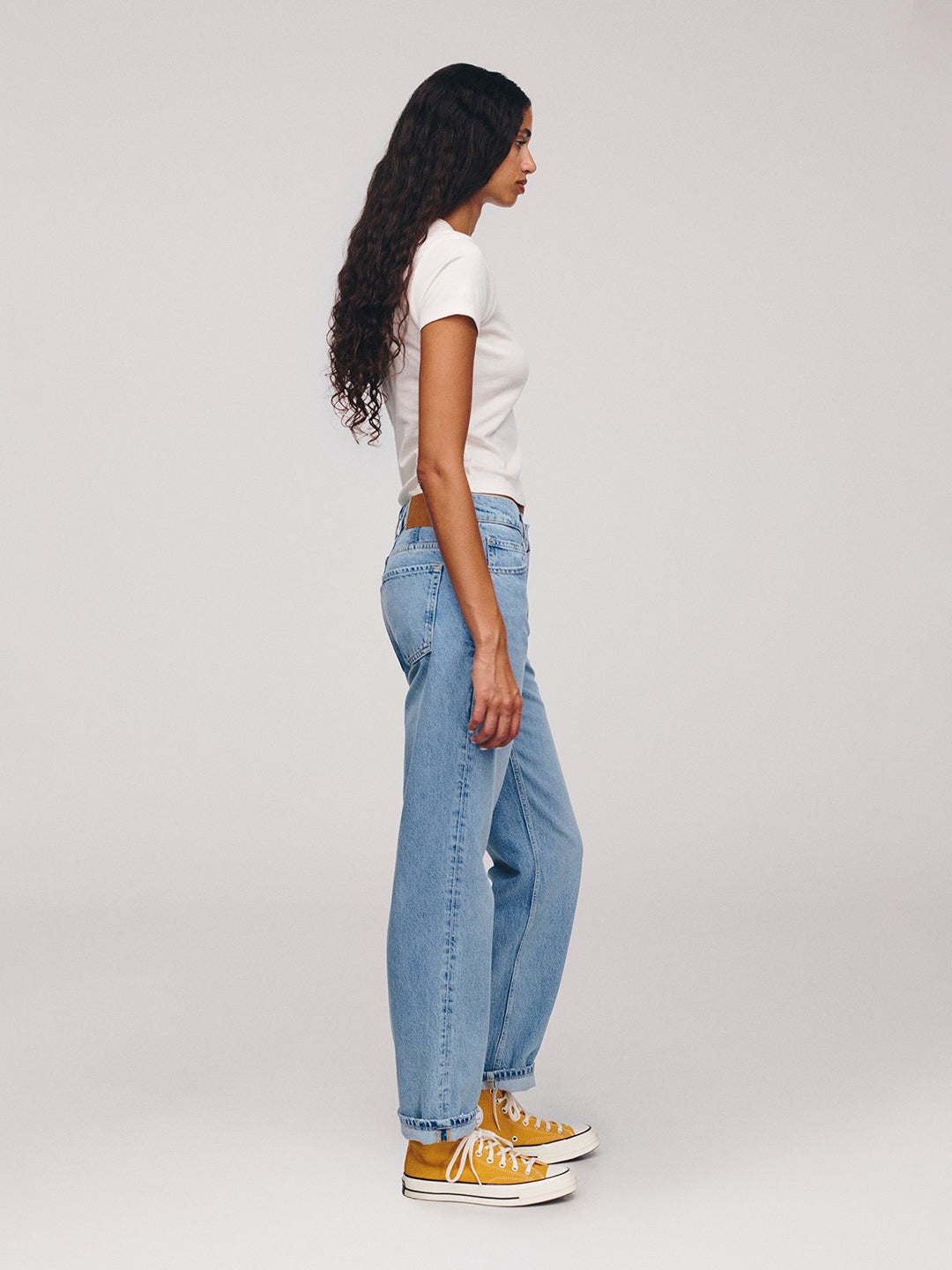 Woman in side profile wearing a white T shirt, relaxed blue jeans, and white high top sneakers against a plain light background
