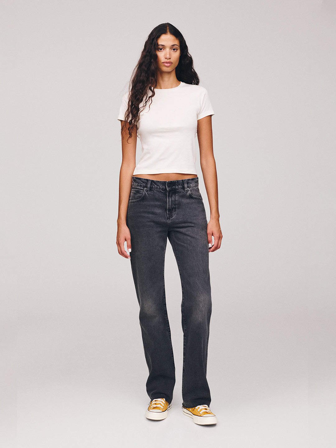 Woman with long dark hair wearing a white fitted T shirt dark straight leg jeans and white sneakers standing against a light gray studio background