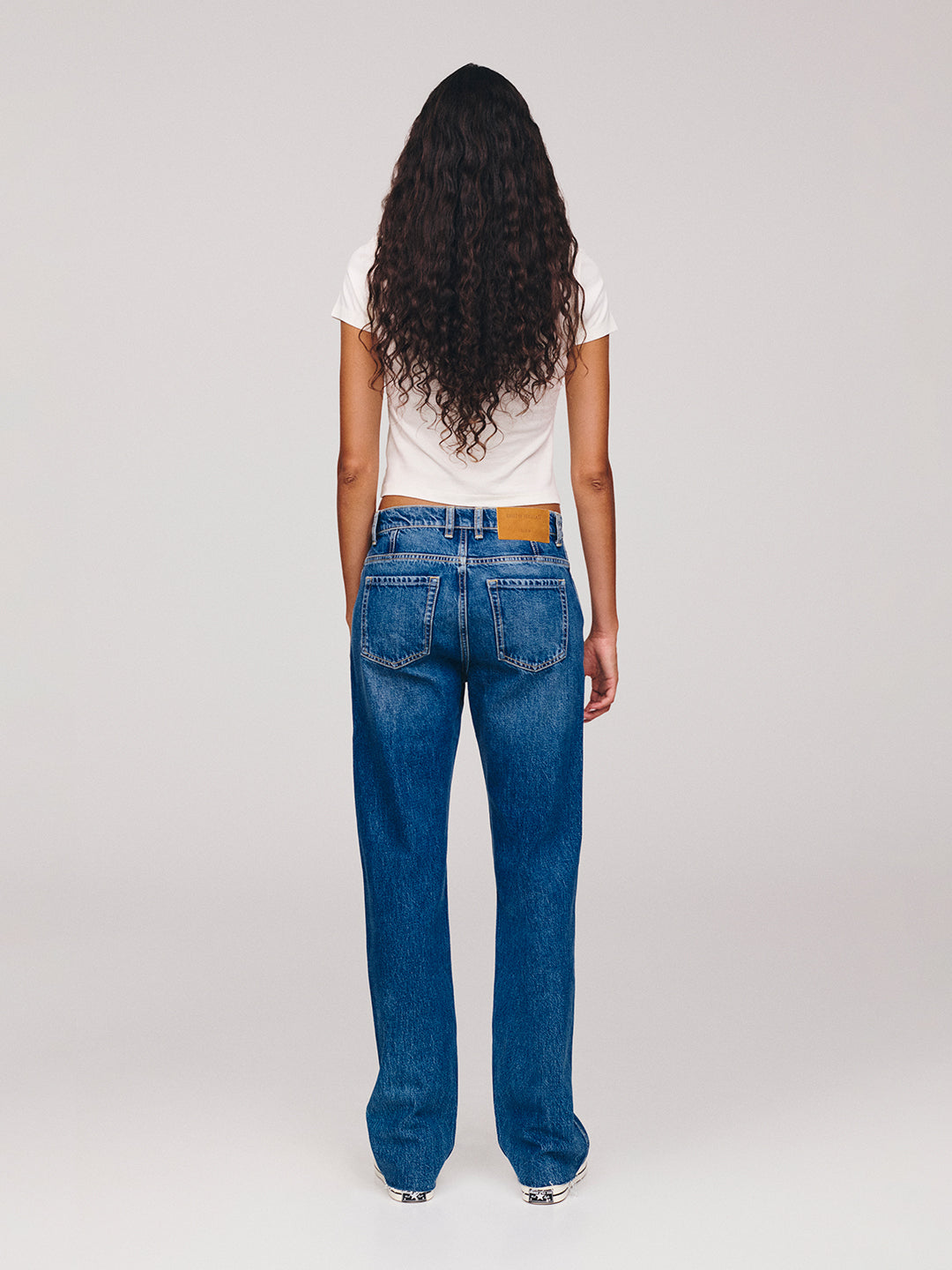 Back view of a woman with long curly hair wearing a white T shirt and relaxed blue jeans against a plain light gray studio background