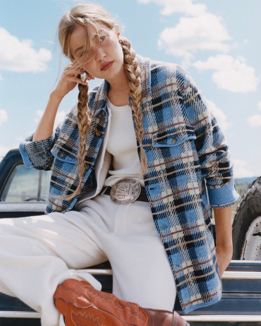 Young woman with braided hair sits on a vintage vehicle wearing a blue plaid jacket over a white outfit beneath a bright blue sky