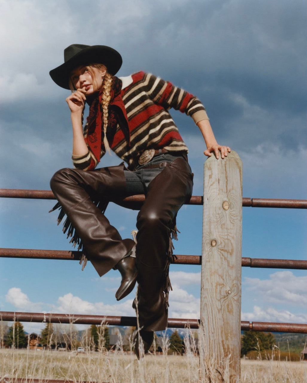 A person in a cowboy hat, striped sweater, fringe leather pants, and boots sits on a metal fence next to a wooden post outdoors under a cloudy sky.