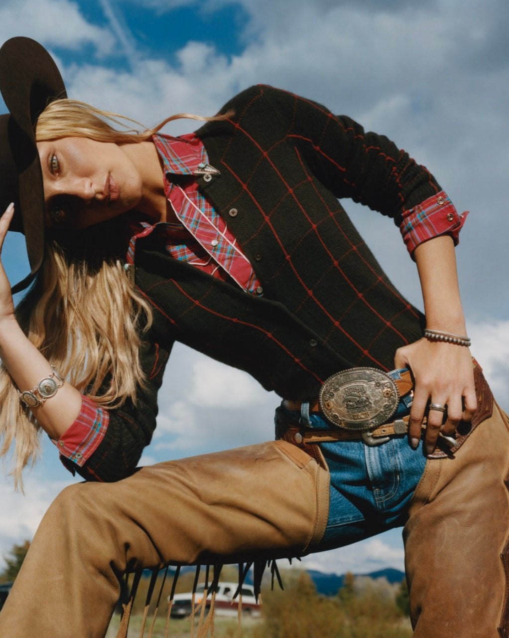 Woman in a cowboy hat wearing a black and red plaid cardigan over a matching shirt with jeans and brown trousers in an outdoor setting under a cloudy sky