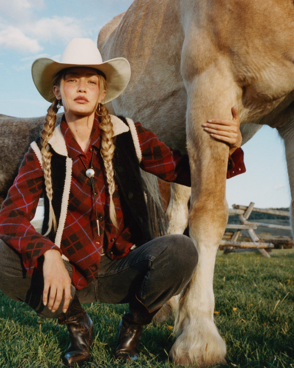Woman in a white cowboy hat red plaid shirt and black vest crouching beside a large horse in a grassy rural field under a blue sky